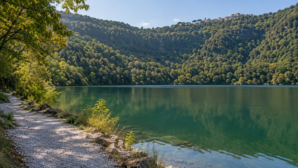 Sentiero di ghiaia lungo la riva del Lago di Nemi, con i boschi del cratere e il borgo sul bordo